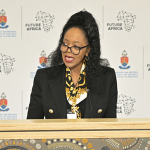 Judy Dlamini looks to her right as she speaks at AACSB's Africa Conference. She has long black shoulder-length hair with a section pulled up in a high pony tail, and she is wearing black round reading glasses, hoop earrings, and a black blazer with one large gold button beside each lapel over a white shirt with an ornate black, gold, and white scarf tied casually around her neck. She stands against a white backdrop with the logos for the University of Pretoria and the Future Africa campus printed repeatedly on it.