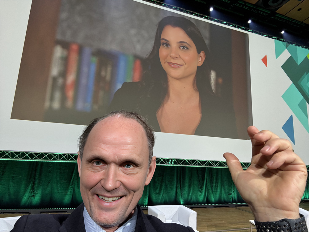 Johan Roos smiles and waves as he takes a selfie of himself in front of a screen that displays Astrid, the AI voice assistant he programmed to interact with participants in his ICAM conference session. Astrid appears on the screen behind Johan as a smiling woman with a light complexion and long wavy dark brown hair, wearing a black blouse with a deep V-neck and sitting before a shelf filled with books.