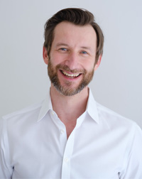 Author and AI entrepreneur Cezary Pietrasik with light complexion, short dark brown hair side parted, blue eyes, very short neatly trimmed brown beard and mustache with strands of gray, and friendly open smile. He wears a plain white button-down shirt with the top two buttons open and stands facing the viewer against a plain gray background.