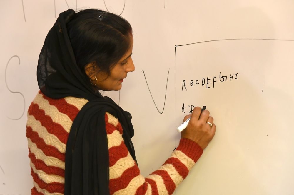 A staff member at BIMTECH, a young woman with long black hair pulled into a ponytail and black scarf over a sweater with wide red-and-white wavy stripes, stands at a whiteboard writing the English alphabet.