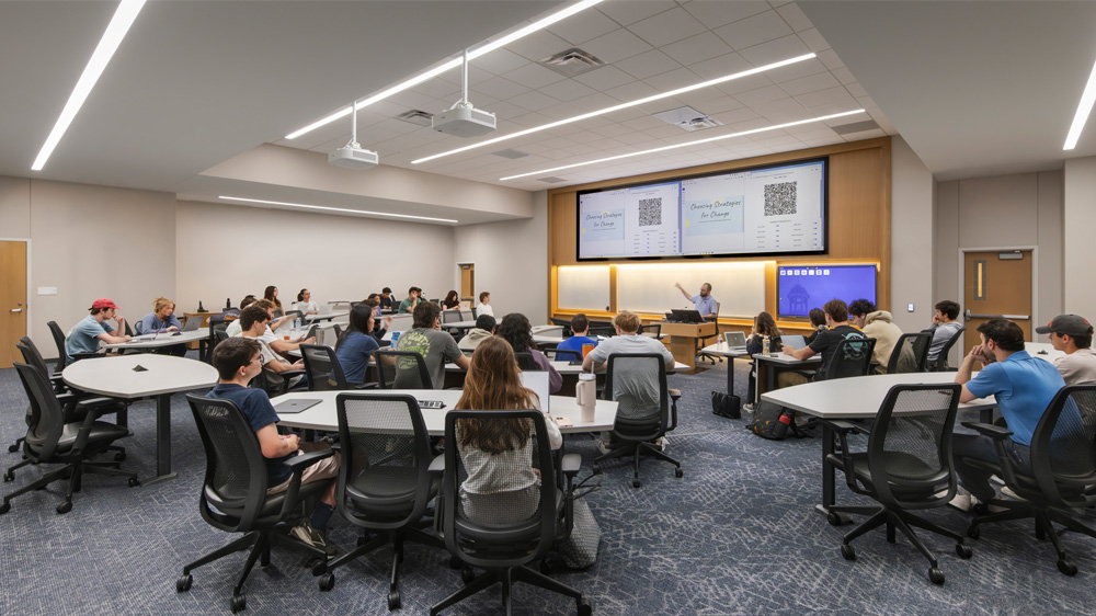 A mid-sized flat-floored gray-carpeted classroom at Southern Methodist University's Cox School of Business with oval-shaped white-topped tables, straight-edged on both short ends, and black mesh-backed rolling chairs, with each table accommodating three to six students for group work. The tables are spaced around the room while a professor presents at the front beneath multiple large display screens, with white projectors installed in the ceiling.